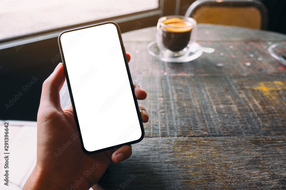 Mockup image of a hand holding and showing black mobile phone with blank white screen and coffee cup on wooden table