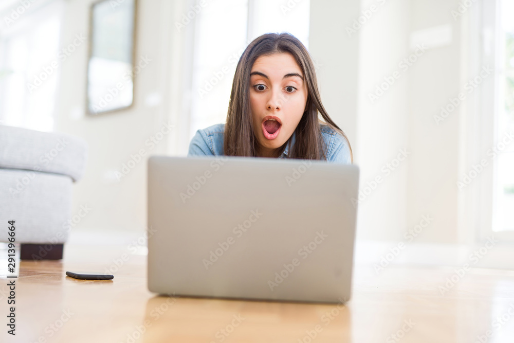 Beautiful young woman laying on the floor using laptop scared in shock with a surprise face, afraid and excited with fear expression Beautiful young woman laying on the floor using laptop scared in shock with a surprise face, afraid and excited with fear expression