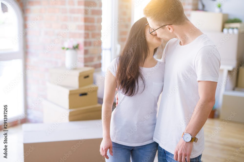 Young beautiful couple kissing wearing glasses standing at new home around cardboard boxes Young beautiful couple kissing wearing glasses standing at new home around cardboard boxes