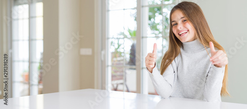 Wide angle picture of beautiful young girl kid wearing casual sweater approving doing positive gesture with hand, thumbs up smiling and happy for success. Looking at the camera, winner gesture.