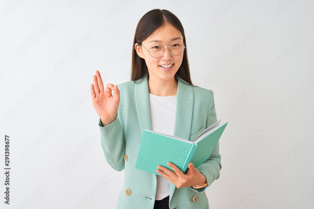 Fototapeta premium Young chinese student woman wearing glasses reading book over isolated white background doing ok sign with fingers, excellent symbol
