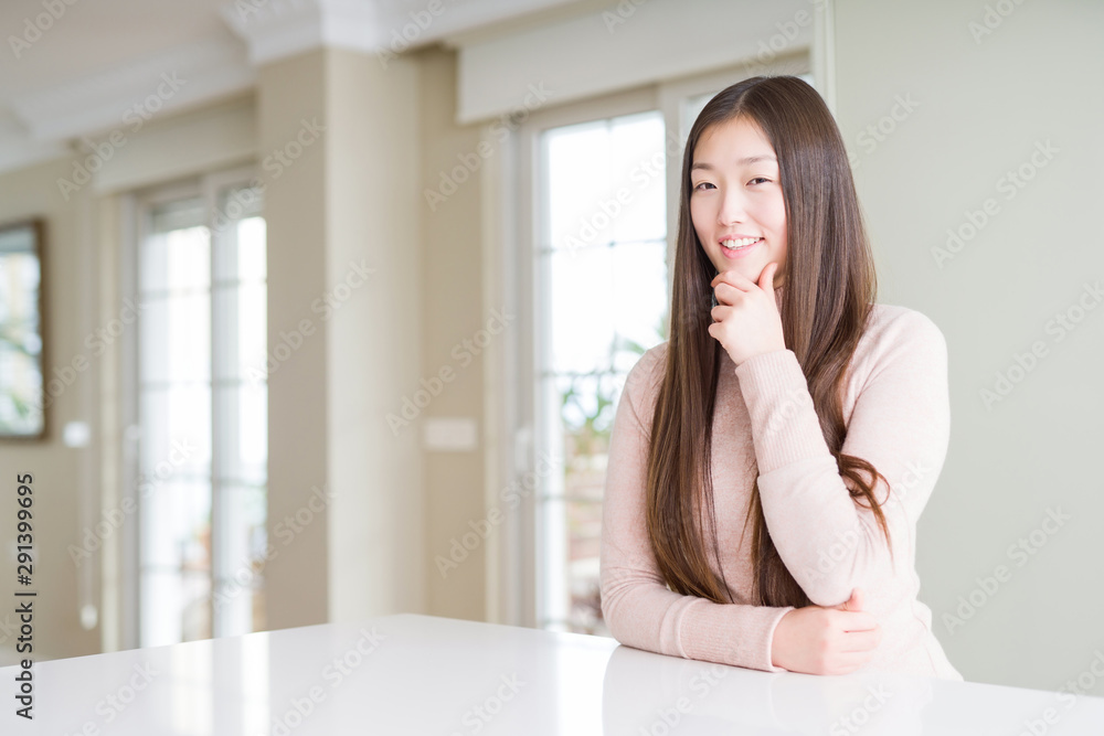 Beautiful Asian woman wearing casual sweater on white table looking confident at the camera with smile with crossed arms and hand raised on chin. Thinking positive. Beautiful Asian woman wearing casual sweater on white table looking confident at the camera with smile with crossed arms and hand raised on chin. Thinking positive.