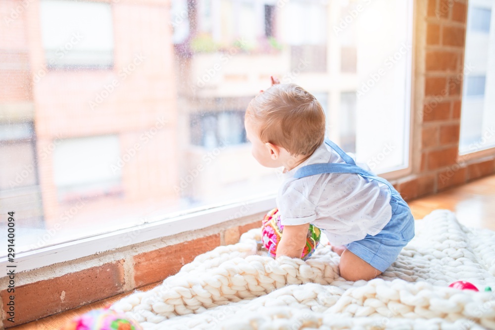 Fototapeta premium Beautiful toddler sitting on the blanket looking through the window at kindergarten
