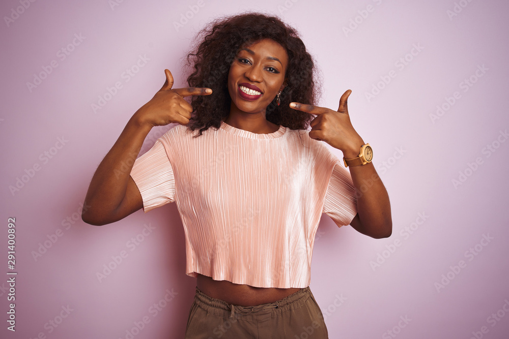 Young african american woman wearing t-shirt standing over isolated pink background smiling cheerful showing and pointing with fingers teeth and mouth. Dental health concept. Young african american woman wearing t-shirt standing over isolated pink background smiling cheerful showing and pointing with fingers teeth and mouth. Dental health concept.