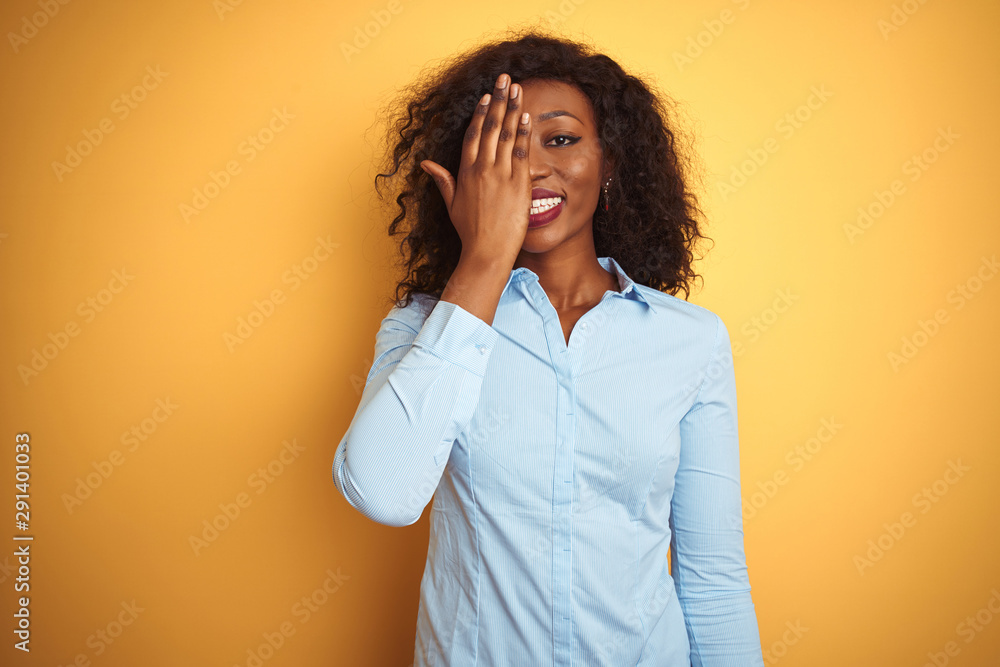 African american businesswoman wearing elegant shirt over isolated yellow background covering one eye with hand, confident smile on face and surprise emotion. African american businesswoman wearing elegant shirt over isolated yellow background covering one eye with hand, confident smile on face and surprise emotion.