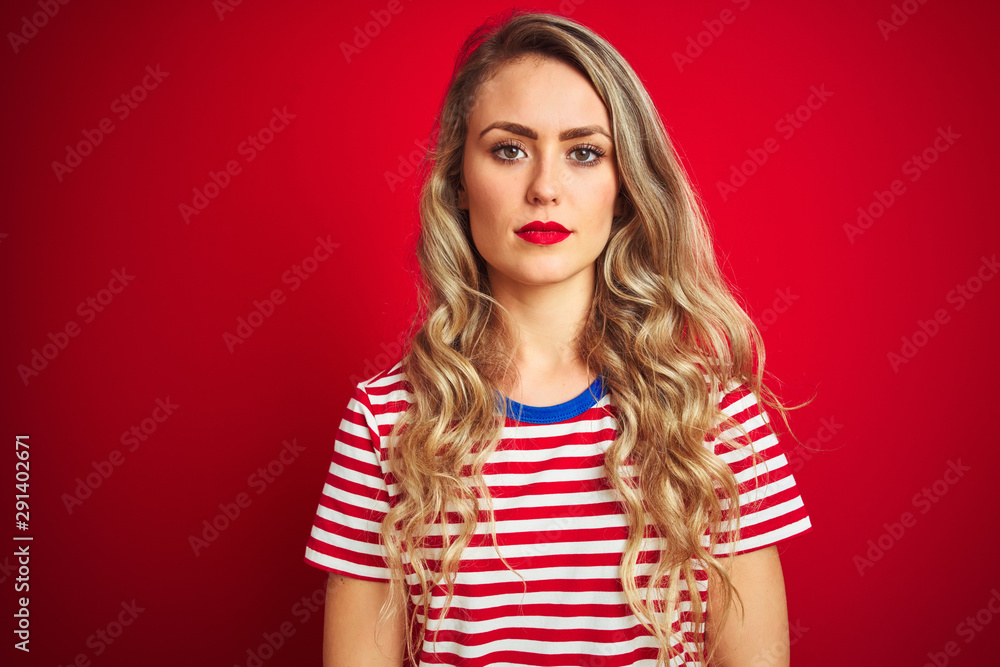 Young beautiful woman wearing stripes t-shirt standing over red isolated background with serious expression on face. Simple and natural looking at the camera. Young beautiful woman wearing stripes t-shirt standing over red isolated background with serious expression on face. Simple and natural looking at the camera.
