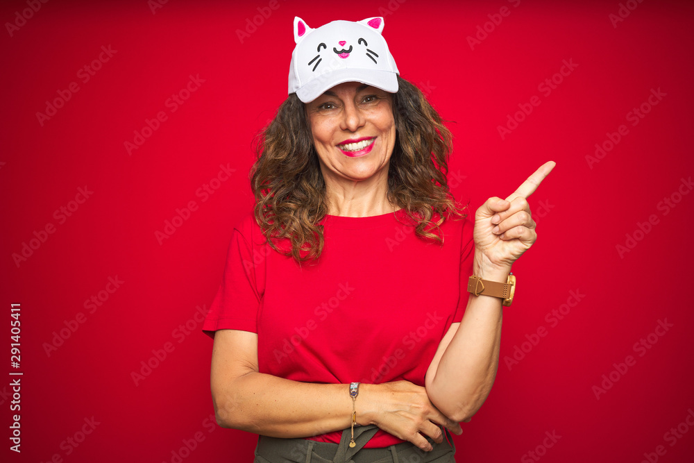 Middle age senior woman wearing cute kitty cap over red isolated background very happy pointing with hand and finger to the side Middle age senior woman wearing cute kitty cap over red isolated background very happy pointing with hand and finger to the side