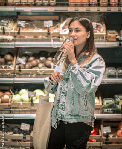 young girl drinking water from a glass bottle with an eco-bag on the background of fruits and vegetables in grociery store photo