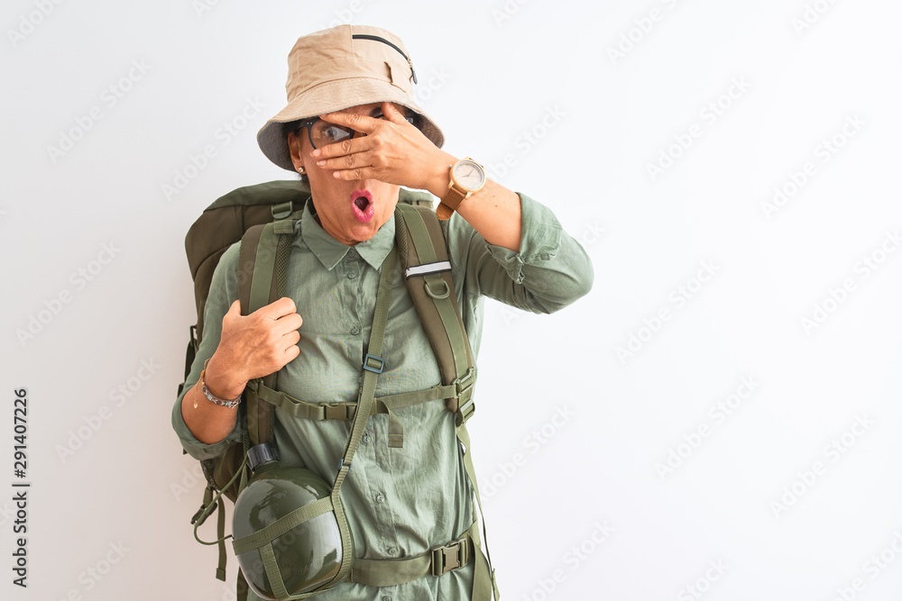 Middle age hiker woman wearing backpack canteen hat glasses over isolated white background peeking in shock covering face and eyes with hand, looking through fingers with embarrassed expression.