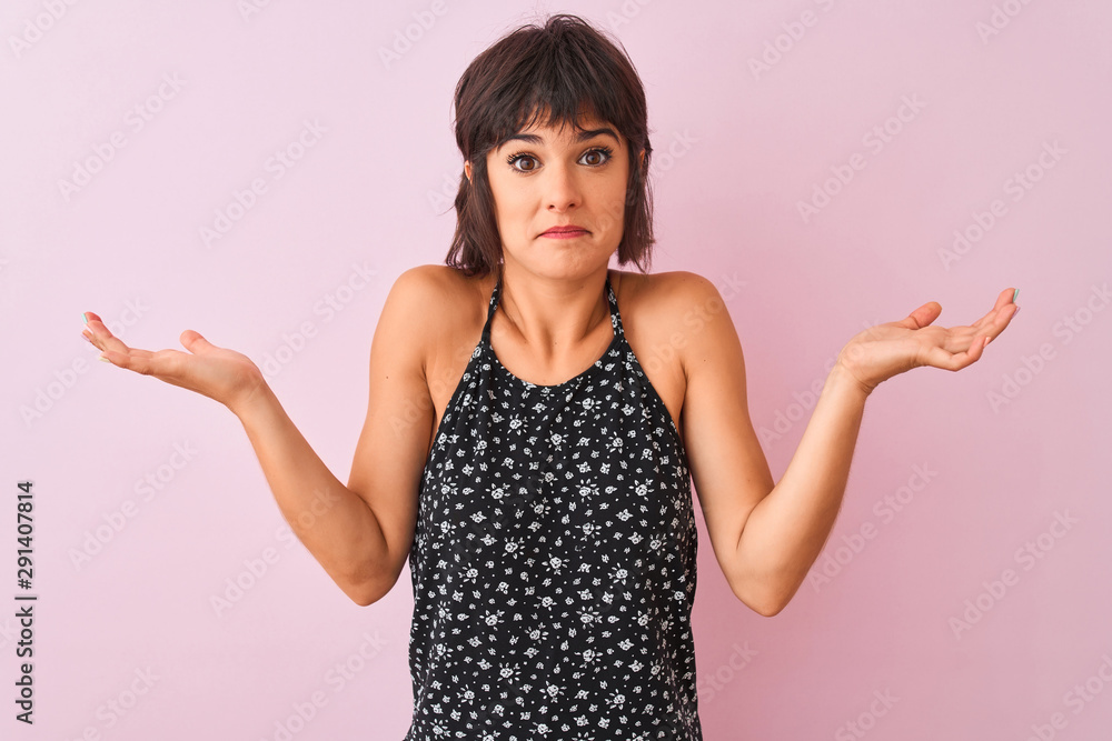 Young beautiful woman wearing black floral dress standing over isolated pink background clueless and confused expression with arms and hands raised. Doubt concept. Young beautiful woman wearing black floral dress standing over isolated pink background clueless and confused expression with arms and hands raised. Doubt concept.