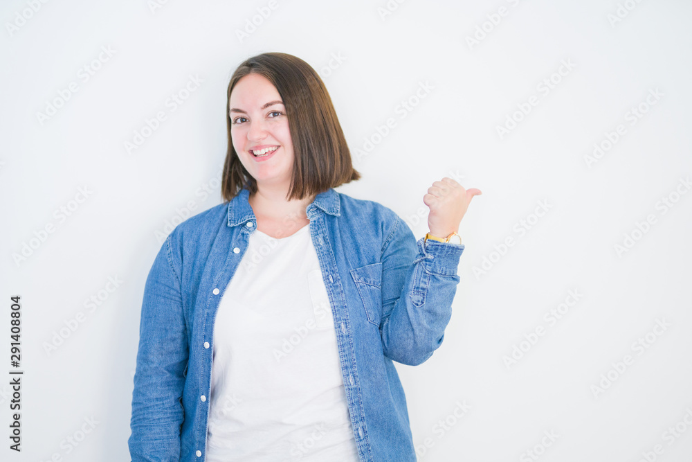 Young beautiful plus size woman over white isolated background smiling with happy face looking and pointing to the side with thumb up. Young beautiful plus size woman over white isolated background smiling with happy face looking and pointing to the side with thumb up.