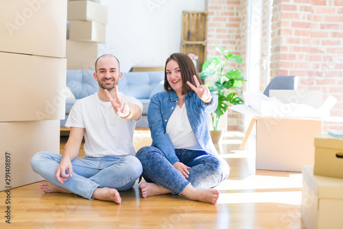 Young couple sitting on the floor arround cardboard boxes moving to a new house smiling with happy face winking at the camera doing victory sign. Number two.