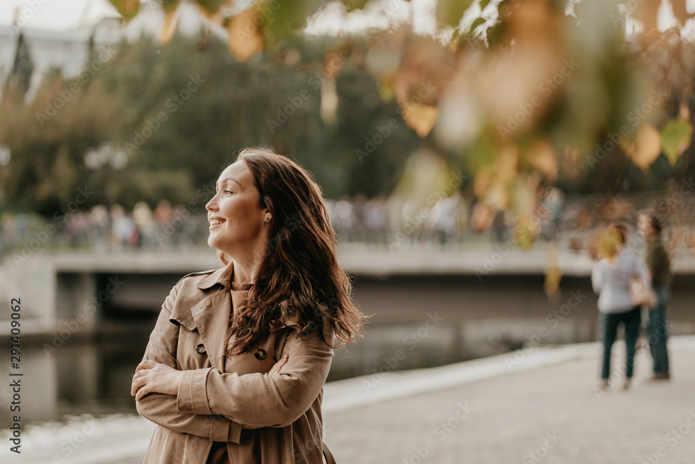 Friendly charming brunette woman with long curly hair in brown coat at street with golden leaves, autumn season Friendly charming brunette woman with long curly hair in brown coat at street with golden leaves, autumn season