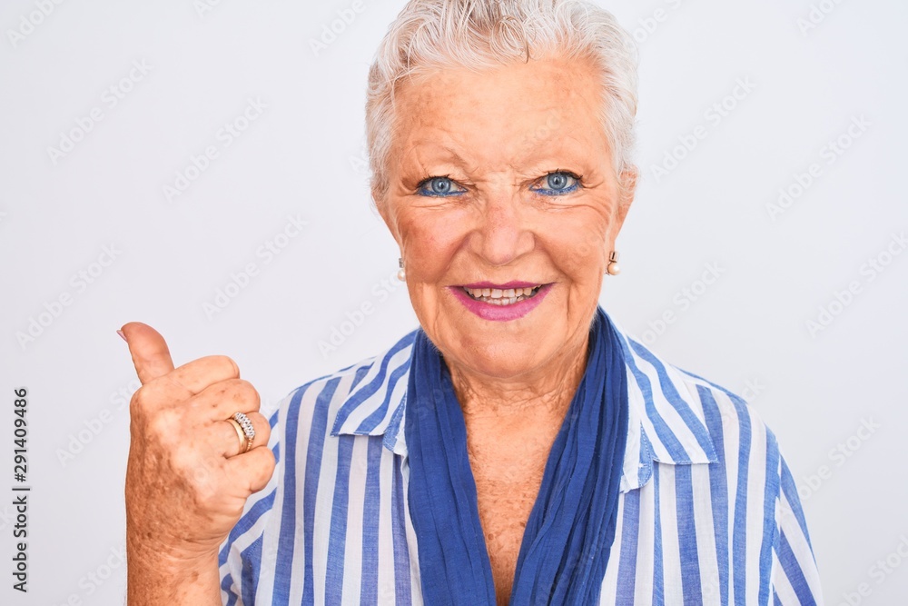 Senior grey-haired woman wearing blue striped shirt standing over isolated white background pointing and showing with thumb up to the side with happy face smiling Senior grey-haired woman wearing blue striped shirt standing over isolated white background pointing and showing with thumb up to the side with happy face smiling