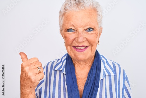 Senior grey-haired woman wearing blue striped shirt standing over isolated white background pointing and showing with thumb up to the side with happy face smiling