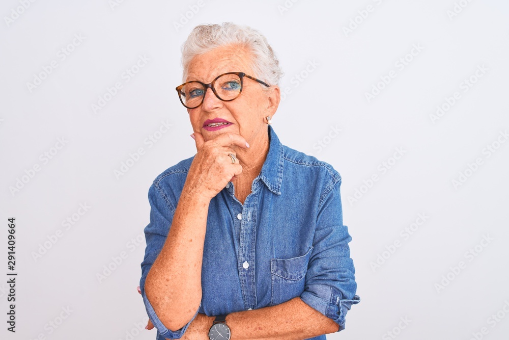 Senior grey-haired woman wearing denim shirt and glasses over isolated white background looking confident at the camera smiling with crossed arms and hand raised on chin. Thinking positive. Senior grey-haired woman wearing denim shirt and glasses over isolated white background looking confident at the camera smiling with crossed arms and hand raised on chin. Thinking positive.
