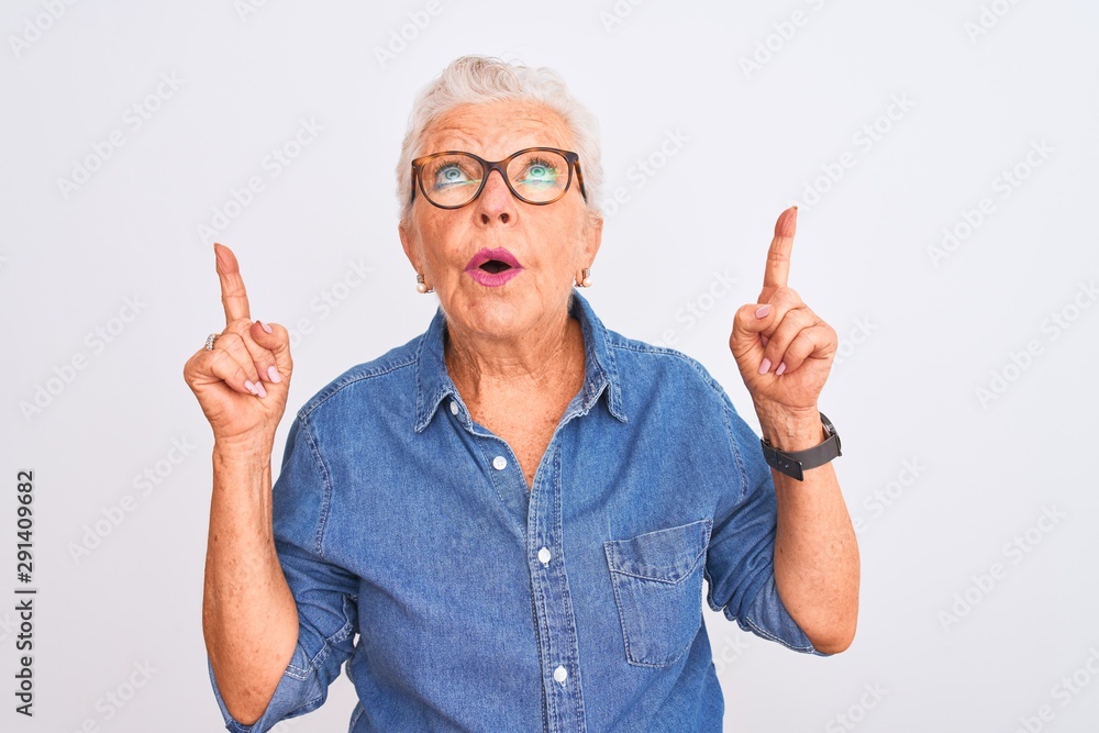 Senior grey-haired woman wearing denim shirt and glasses over isolated white background amazed and surprised looking up and pointing with fingers and raised arms. Senior grey-haired woman wearing denim shirt and glasses over isolated white background amazed and surprised looking up and pointing with fingers and raised arms.