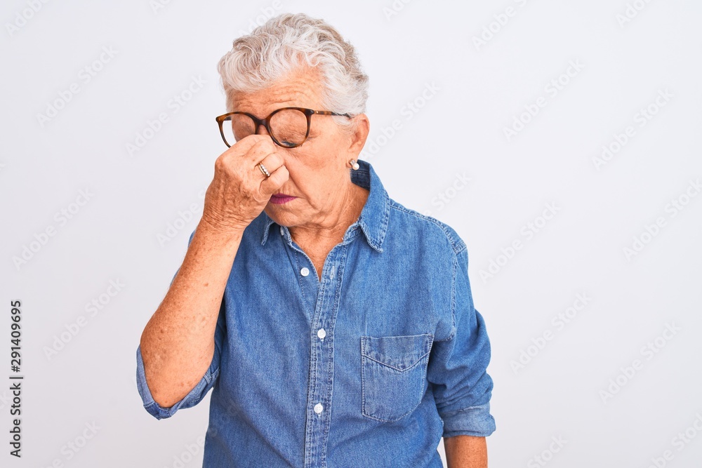 Senior grey-haired woman wearing denim shirt and glasses over isolated white background tired rubbing nose and eyes feeling fatigue and headache. Stress and frustration concept. Senior grey-haired woman wearing denim shirt and glasses over isolated white background tired rubbing nose and eyes feeling fatigue and headache. Stress and frustration concept.