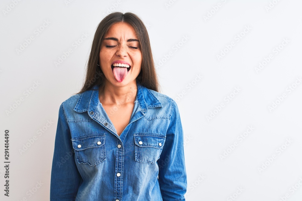 Young beautiful woman wearing casual denim shirt standing over isolated white background sticking tongue out happy with funny expression. Emotion concept. Young beautiful woman wearing casual denim shirt standing over isolated white background sticking tongue out happy with funny expression. Emotion concept.