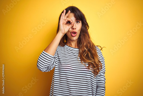 Young beautiful woman wearing stripes t-shirt standing over yelllow isolated background doing ok gesture shocked with surprised face, eye looking through fingers. Unbelieving expression.