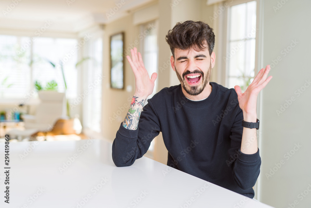 Young man wearing casual sweatshirt sitting on white table celebrating mad and crazy for success with arms raised and closed eyes screaming excited. Winner concept Young man wearing casual sweatshirt sitting on white table celebrating mad and crazy for success with arms raised and closed eyes screaming excited. Winner concept