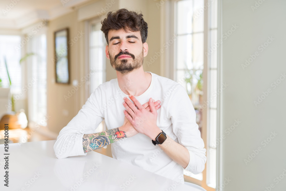 Young man wearing casual shirt sitting on white table smiling with hands on chest with closed eyes and grateful gesture on face. Health concept.