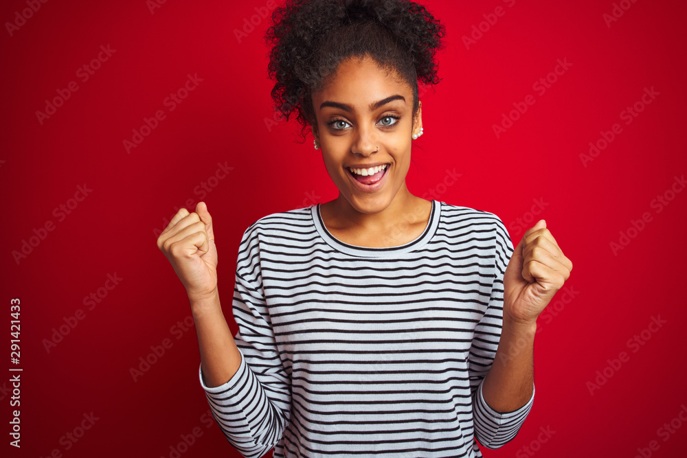 African american woman wearing navy striped t-shirt standing over isolated red background celebrating surprised and amazed for success with arms raised and open eyes. Winner concept. African american woman wearing navy striped t-shirt standing over isolated red background celebrating surprised and amazed for success with arms raised and open eyes. Winner concept.