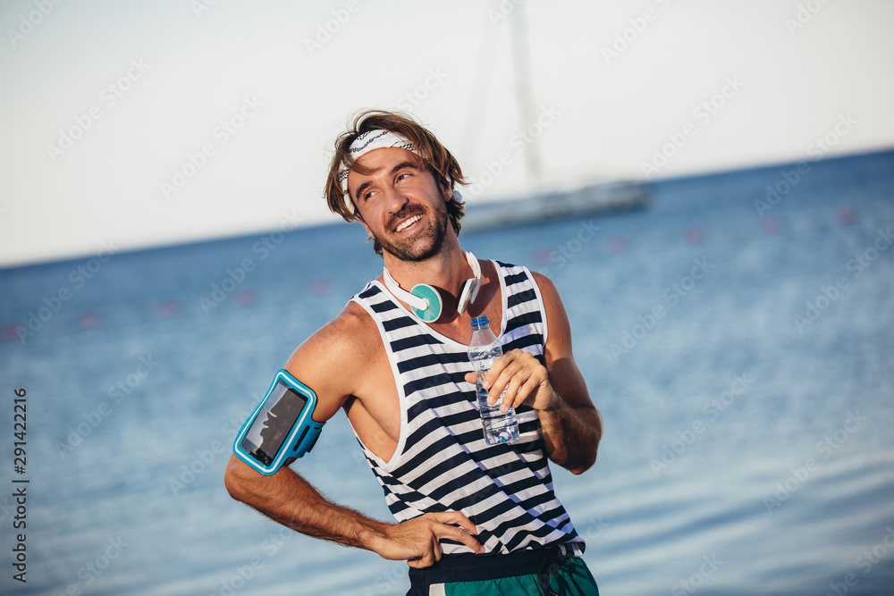 Athletic man with fit body holding bottle of refreshing water, resting after workout or running at beach.