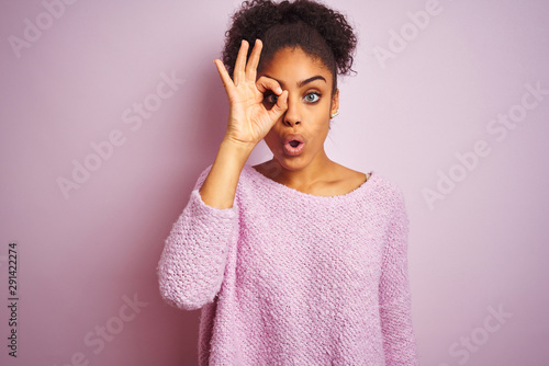 Young african american woman wearing winter sweater standing over isolated pink background doing ok gesture shocked with surprised face, eye looking through fingers. Unbelieving expression.