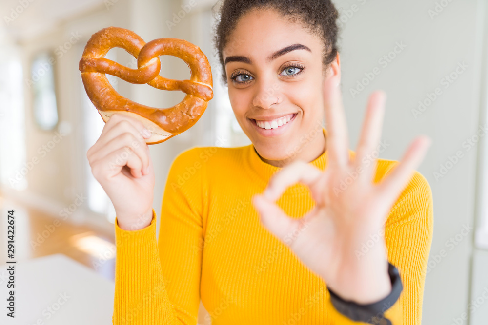 Young african american woman eating German salty pastry pretzel doing ok sign with fingers, excellent symbol