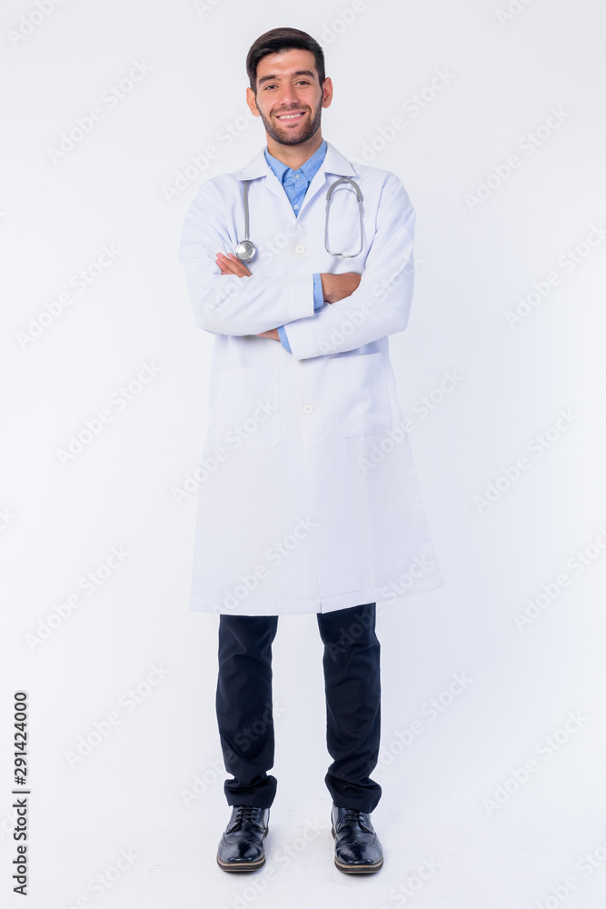 Full body shot of happy young bearded Persian man doctor smiling with arms crossed