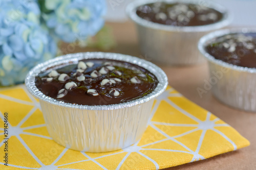 Dark chocolate cake with chocolate chips topping in round foil cup on yellow tablecloth and wooden table. Copy space for your text. Selective focus.