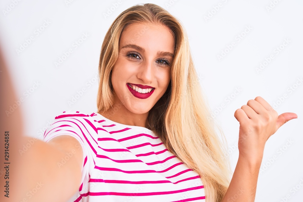 Beautiful woman wearing striped t-shirt make selfie by camera over isolated white background pointing and showing with thumb up to the side with happy face smiling Beautiful woman wearing striped t-shirt make selfie by camera over isolated white background pointing and showing with thumb up to the side with happy face smiling
