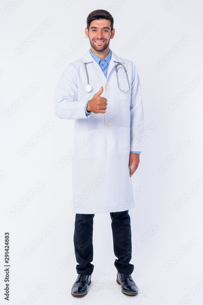 Full body shot of happy young bearded Persian man doctor giving thumbs up Full body shot of happy young bearded Persian man doctor giving thumbs up