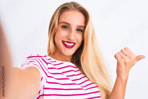Beautiful woman wearing striped t-shirt make selfie by camera over isolated white background pointing and showing with thumb up to the side with happy face smiling