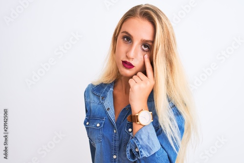 Young beautiful woman wearing casual denim shirt standing over isolated white background Pointing to the eye watching you gesture, suspicious expression