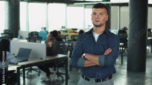 Wallpaper Mural Young businessman standing in a modern office, serious look at the camera, office workers on blurred background. Torontodigital.ca
