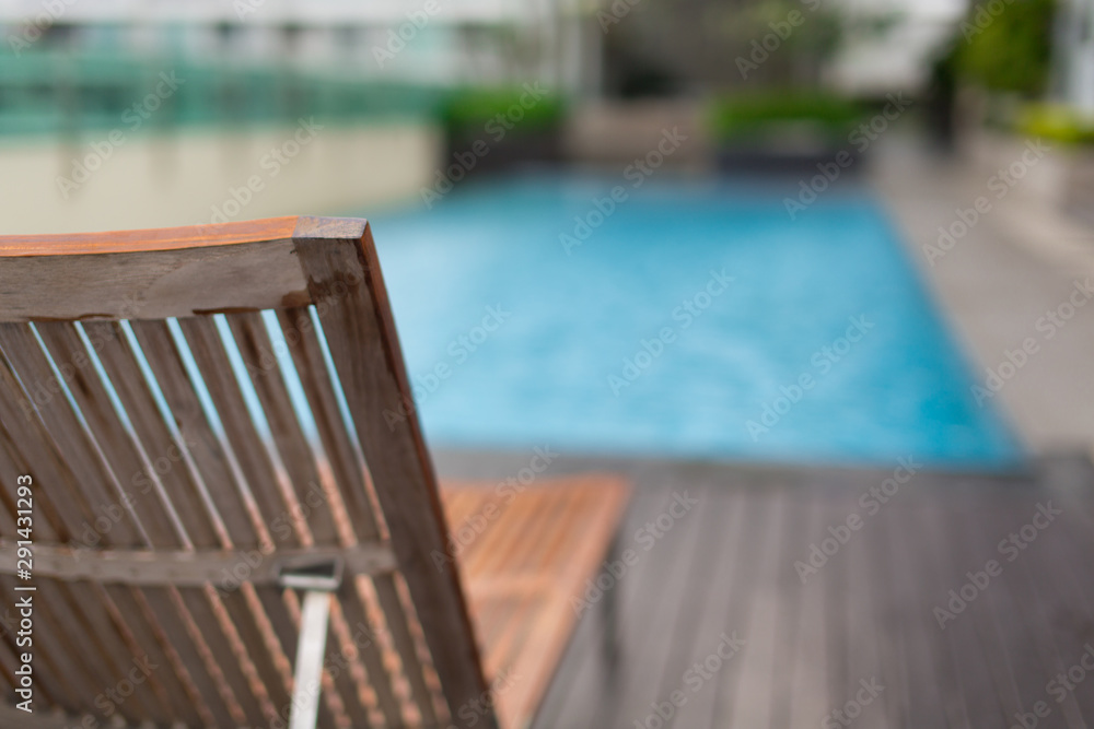 Wooden swimming pool lounger besides a pool with blurred background in sunny day,Thailand. Horizontal shot.