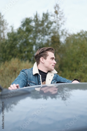 a handsome young man stands behind a car, leans on the roof of a gray car. © Nycowl