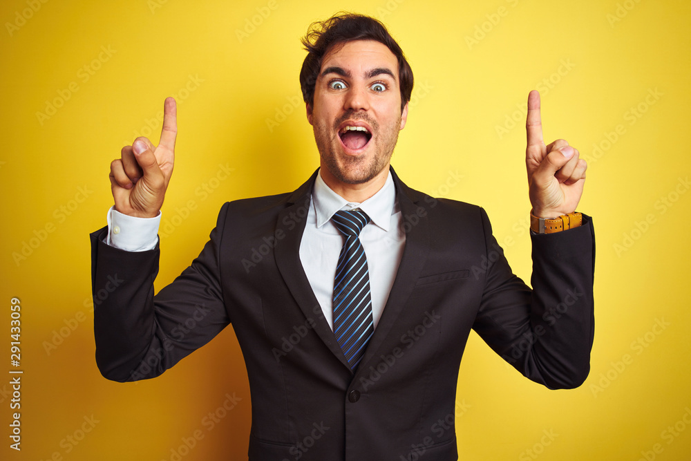 Young handsome businessman wearing suit and tie standing over isolated yellow background smiling amazed and surprised and pointing up with fingers and raised arms. Young handsome businessman wearing suit and tie standing over isolated yellow background smiling amazed and surprised and pointing up with fingers and raised arms.