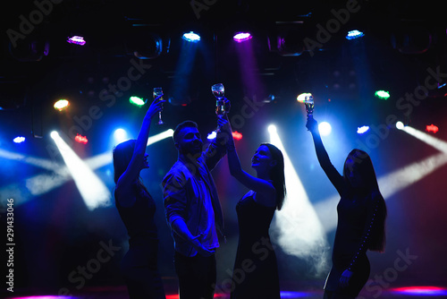 Fototapeta Naklejka Na Ścianę i Meble -   Young friends dancing with glasses of champagne in hands. Against lighting devices as background. Young people's friends are dancing.