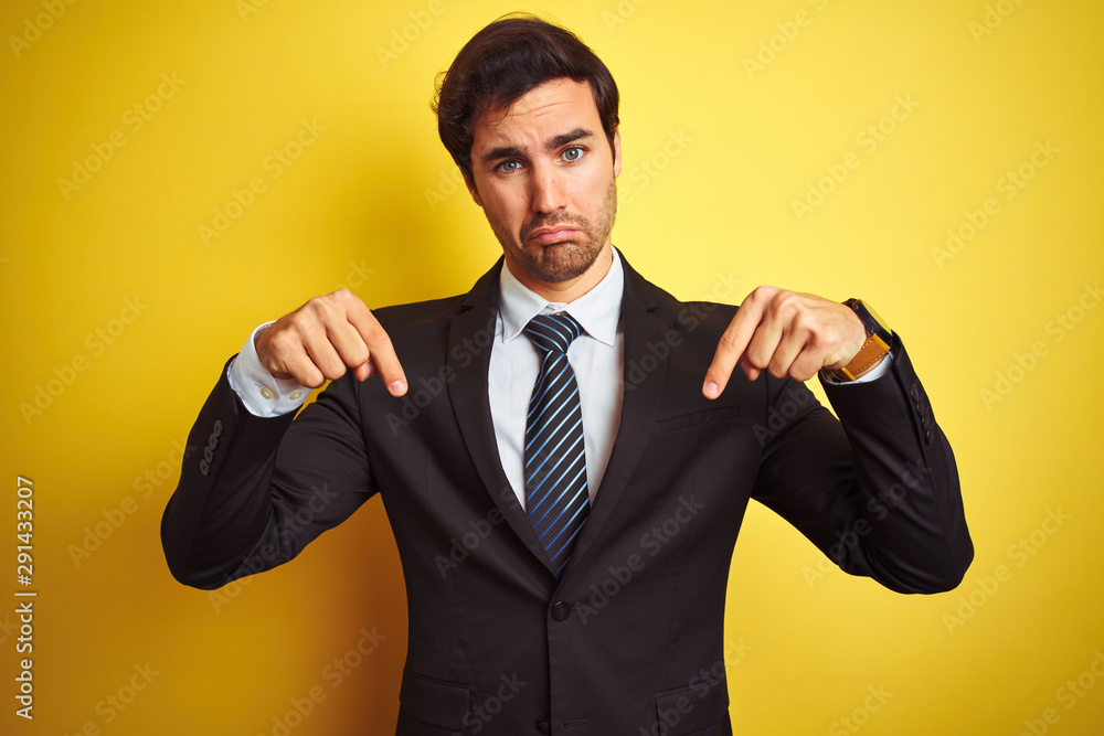 Young handsome businessman wearing suit and tie standing over isolated yellow background Pointing down looking sad and upset, indicating direction with fingers, unhappy and depressed. Young handsome businessman wearing suit and tie standing over isolated yellow background Pointing down looking sad and upset, indicating direction with fingers, unhappy and depressed.