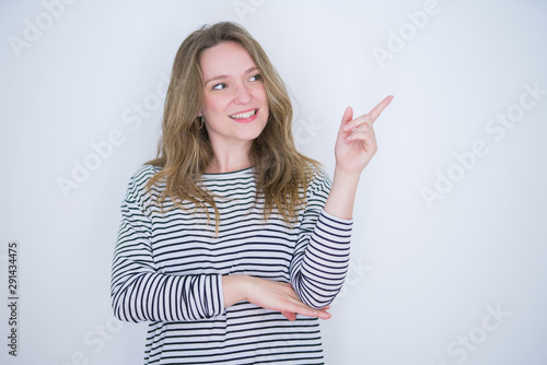 Beautiful blonde girl with blue eyes wearing striped sweater over white isolated background with a big smile on face, pointing with hand and finger to the side looking at the camera.