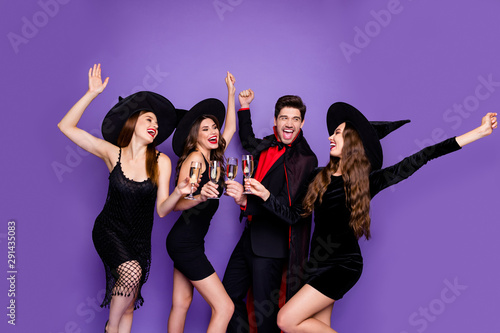 Photo of witch ladies and warlock guy chilling at halloween party drink golden wine raising arms up wear black dresses hats and vampire coat isolated purple color background