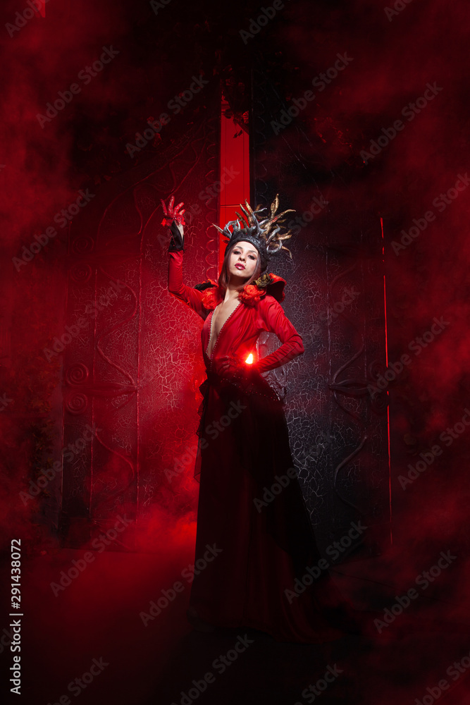 beautiful fashion witch woman with horns in the form of tree roots in a long luxurious dress on the background of a huge gate with red smoke. Halloween concept beautiful fashion witch woman with horns in the form of tree roots in a long luxurious dress on the background of a huge gate with red smoke. Halloween concept