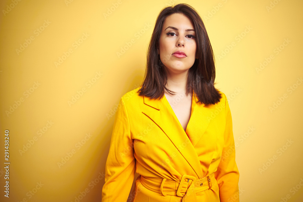 Young beautiful brunette woman wearing elegant dress over yellow isolated background with serious expression on face. Simple and natural looking at the camera. Young beautiful brunette woman wearing elegant dress over yellow isolated background with serious expression on face. Simple and natural looking at the camera.