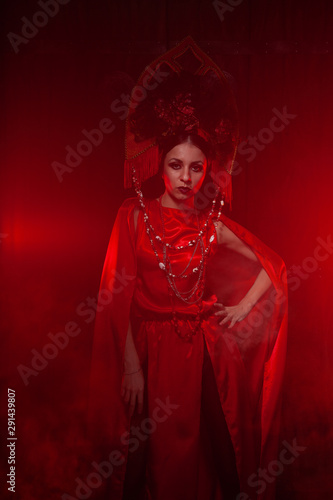 strict and scary girl in a fashionable long red dress and a voluminous hat on the background of smoke in the Studio at night alone. Halloween concept.