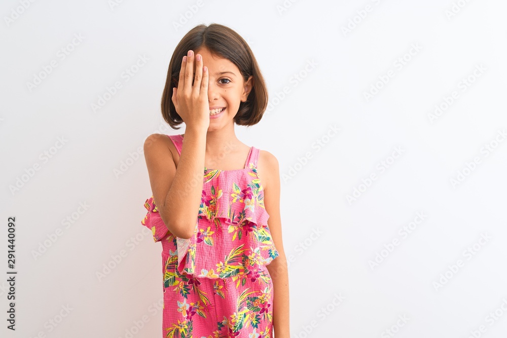 Young beautiful child girl wearing pink floral dress standing over isolated white background covering one eye with hand, confident smile on face and surprise emotion. Young beautiful child girl wearing pink floral dress standing over isolated white background covering one eye with hand, confident smile on face and surprise emotion.
