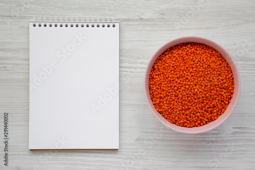 Red lentils in a pink bowl, blank notebook on a white wooden background, top view. Flat lay, overhead, from above. Copy space.
