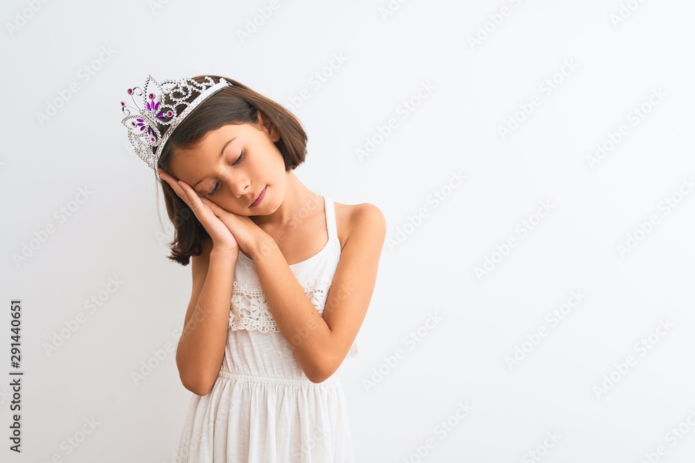 Beautiful child girl wearing princess crown standing over isolated white background sleeping tired dreaming and posing with hands together while smiling with closed eyes. Beautiful child girl wearing princess crown standing over isolated white background sleeping tired dreaming and posing with hands together while smiling with closed eyes.